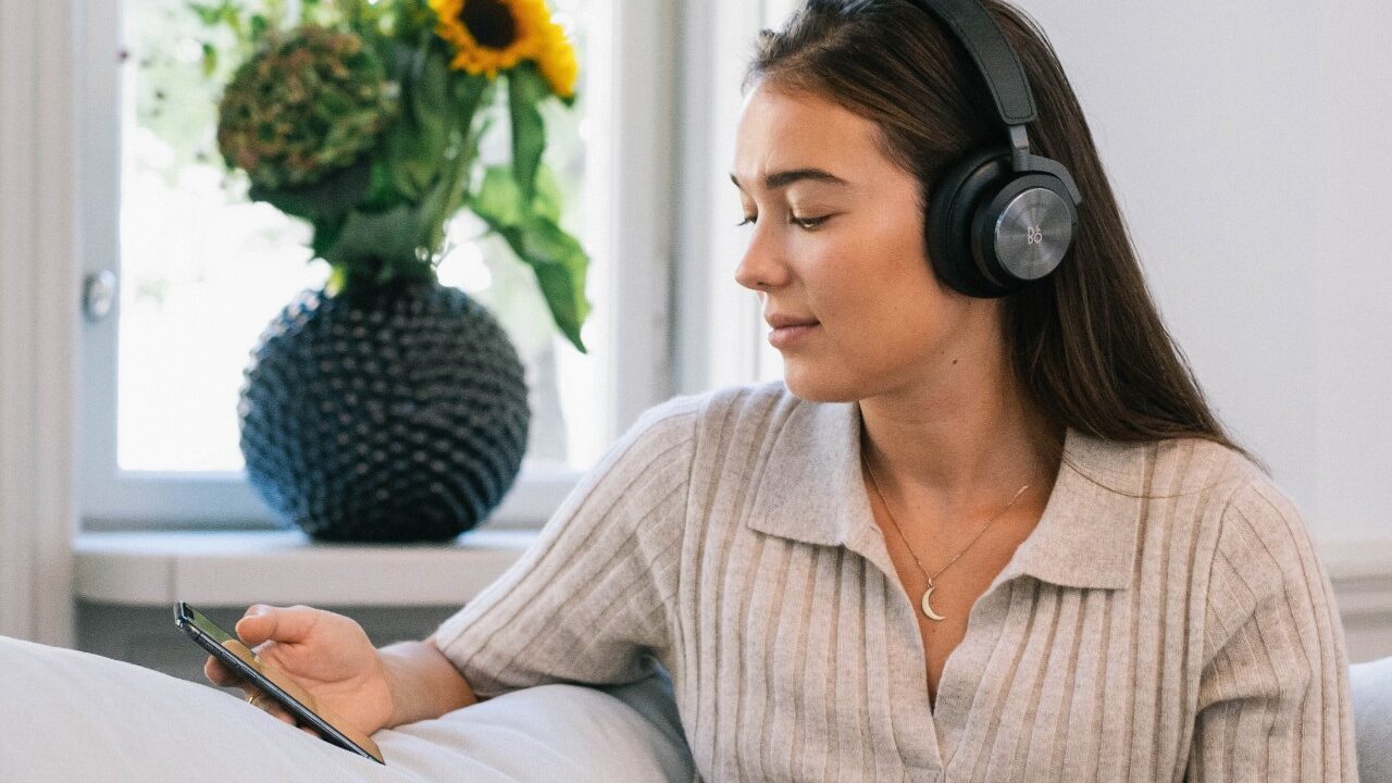 kvinna sitter i soffa med app i handen och hörlurar på sig, med en blomma i bakgrunden